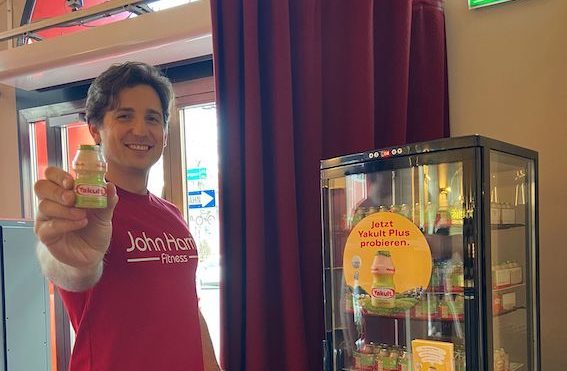 Mann in rotem T-Shirt hält eine Yakult Flasche neben einem Kühlschrank